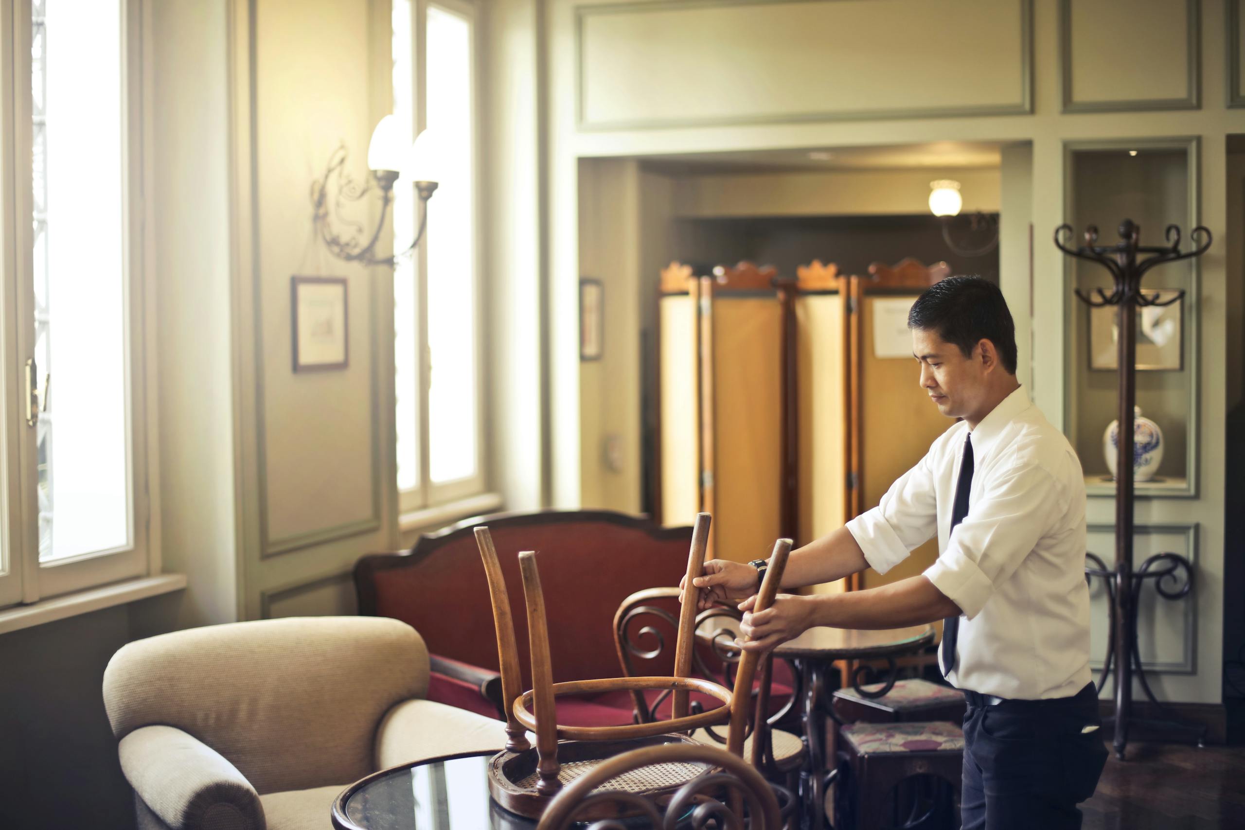 Side view of Asian male employee in formal wear standing near table and neatly stacking wooden chair on table in closed restaurant